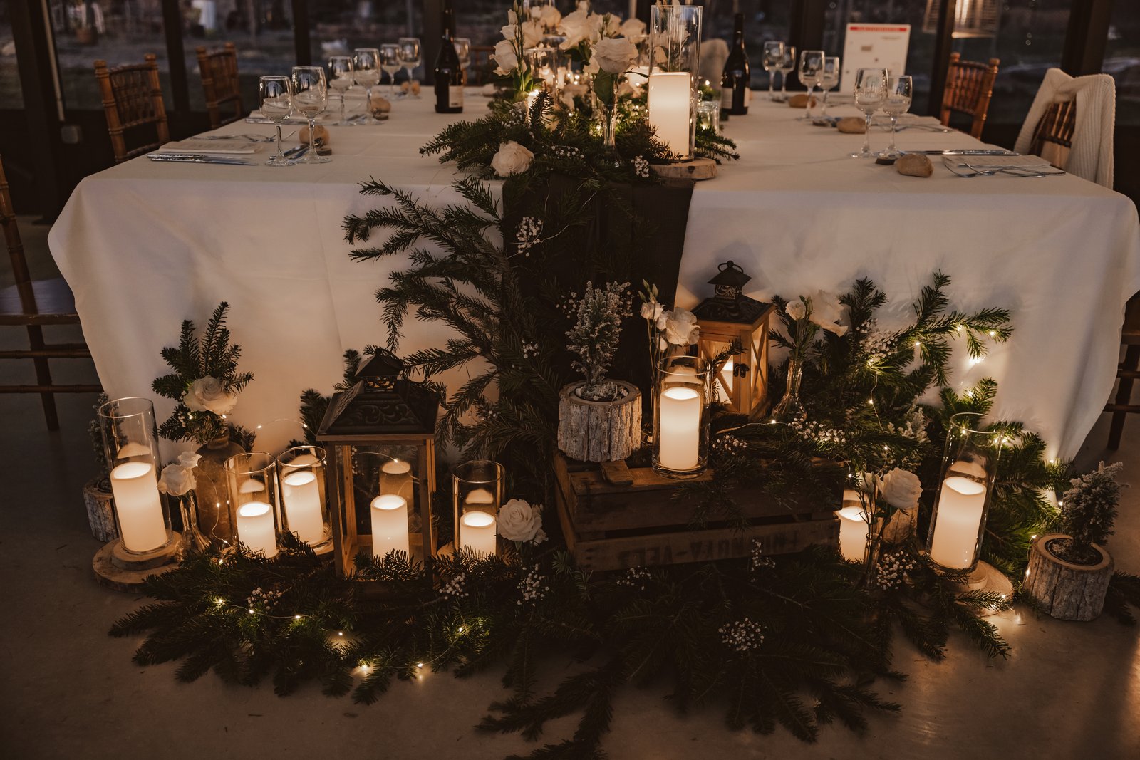 Table d’honneur nappée de blanc, branches de sapin au sol, lanternes en bois, caisse en bois et bougies dans des cylindres de verre