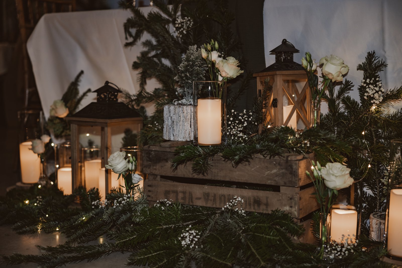 Caisse en bois sur des branches de sapin, lanternes en bois, roses blanches, gypsophile et bougies dans des cylindres de verre