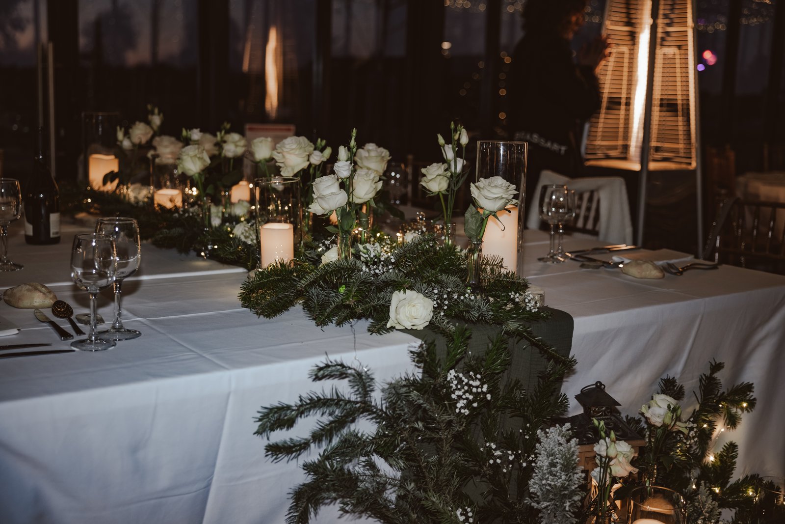 Chemin de sapin, roses blanches en soliflore et bougies sur une table nappée de blanc, chauffage d’extérieur en fond