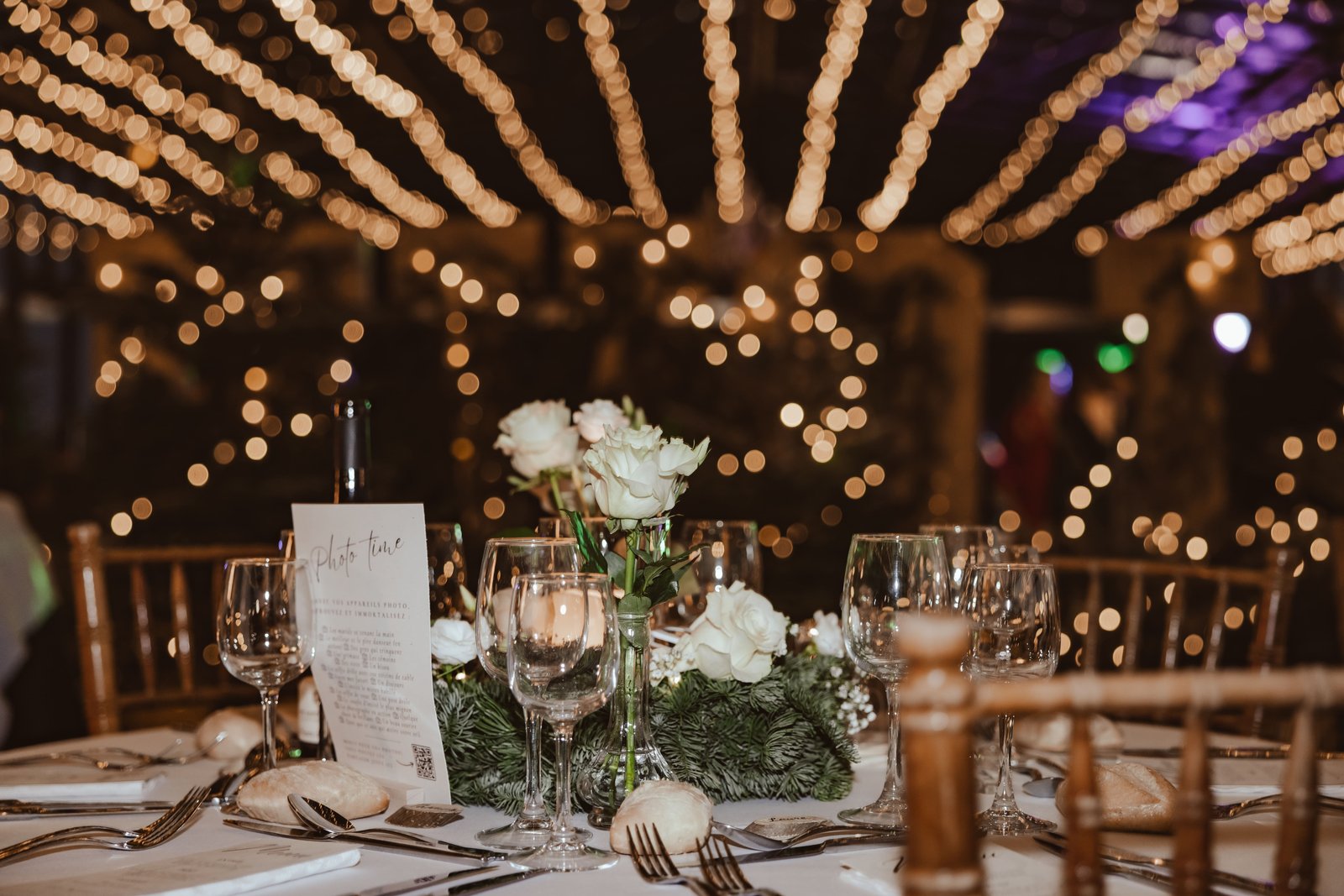 Table ronde dressée de verres et de roses blanches sur couronne de sapin, guirlandes lumineuses floues au plafond