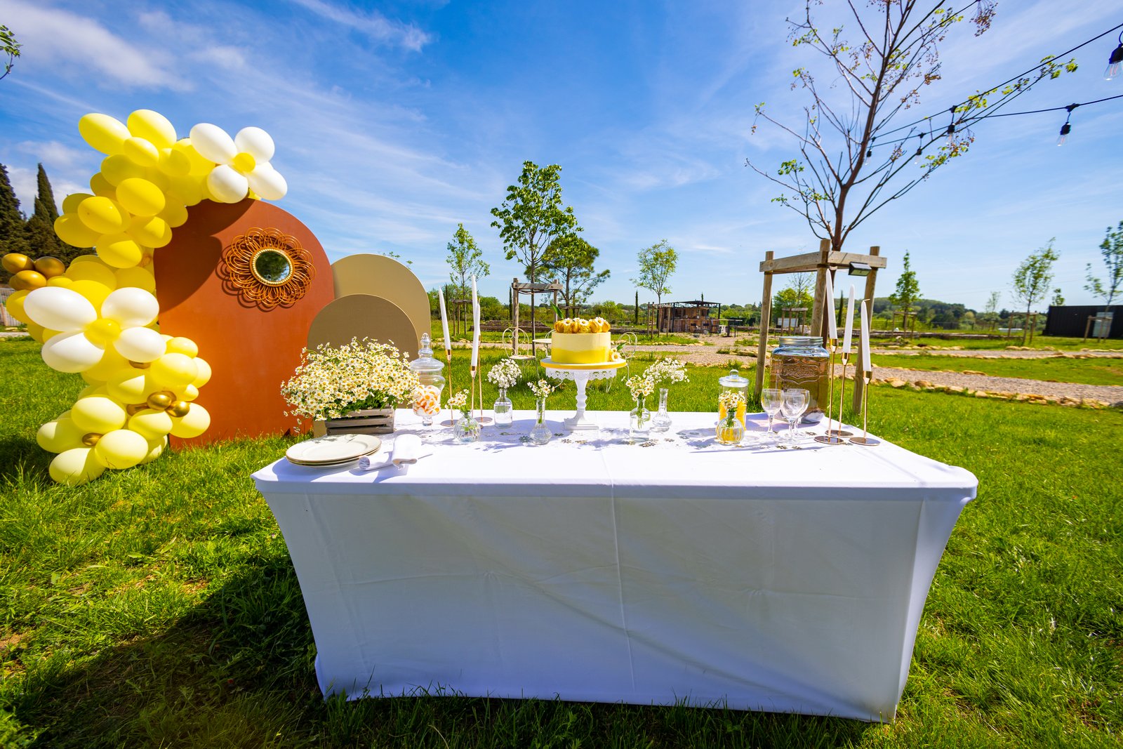 Table nappée de blanc dans un jardin, arches terracotta et sable, guirlande de ballons jaunes et blancs, gâteau jaune