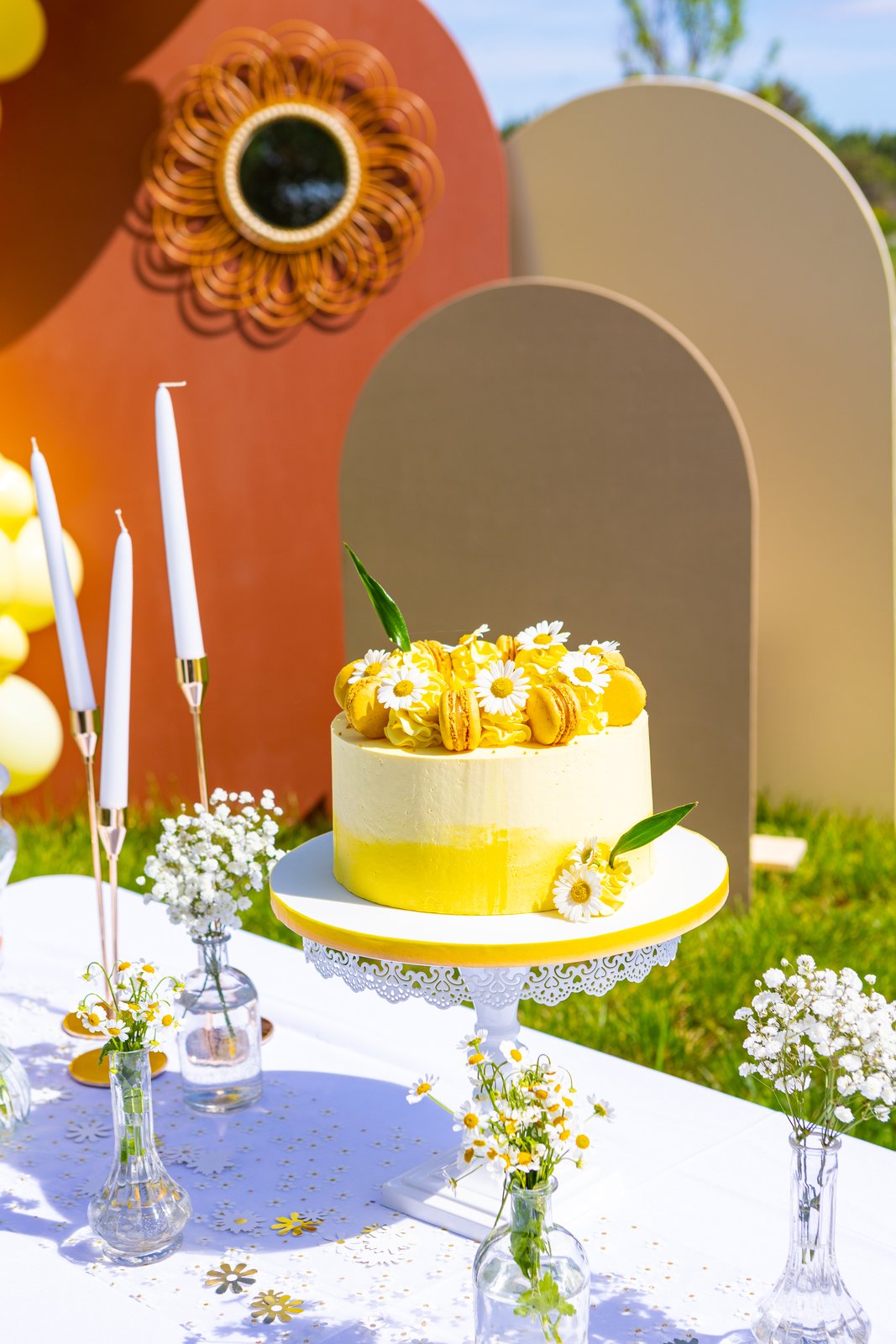 Gâteau jaune aux marguerites et macarons, fines bougies blanches sur bougeoirs dorés, miroir fleur en rotin derrière