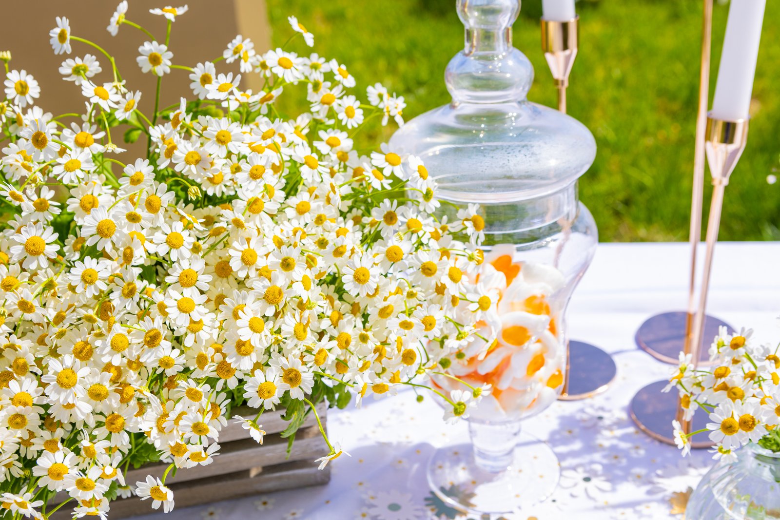 Cagette en bois débordant de marguerites, bonbonnière en verre garnie de bonbons et fins bougeoirs dorés sur nappe blanche