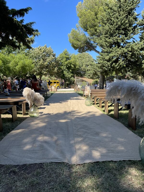 Allée de cérémonie en toile de jute entre des bancs en bois, bordée de dames-jeannes garnies de pampas, sous les grands arbres du domaine