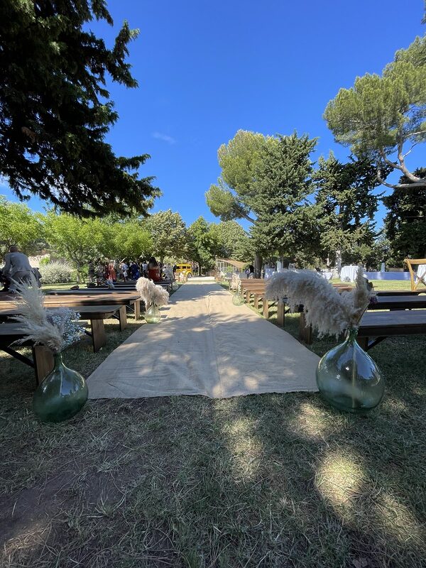 Allée de jute vue depuis l’entrée, dames-jeannes aux pampas et rangs de bancs en bois sous un ciel bleu