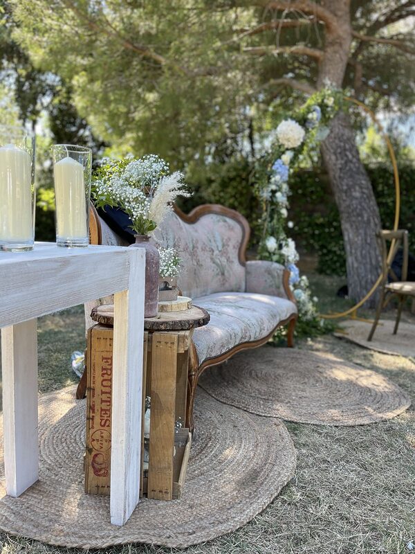 Canapé vintage à motifs fleuris sur des tapis ronds en jute, console blanche aux bougies sous cylindres de verre, caisse en bois et rondin, arche dorée en fond