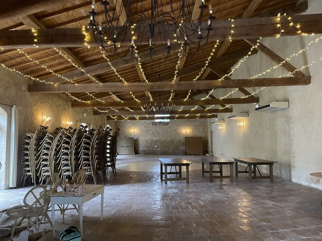 Salle sous charpente en bois traversée de guirlandes LED, lustres en fer forgé et murs de pierre, avant la mise en place
