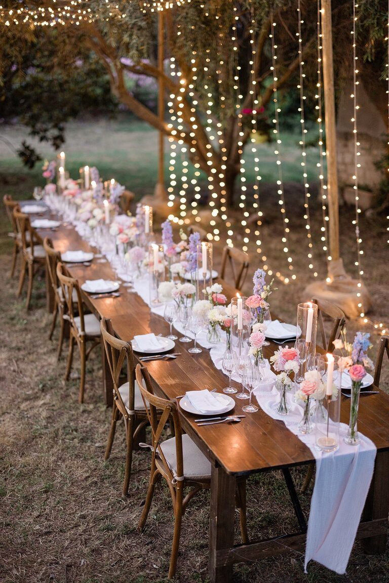 Longue tablée en bois sous un olivier, chaises crossback, chemin de gaze blanche, fleurs pastel et rideaux de leds