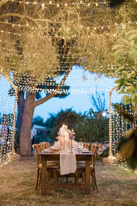 Table en bois dressée sous un ciel étoilé de guirlandes tendu entre des mâts en bois, bougies et fleurs pastel au crépuscule