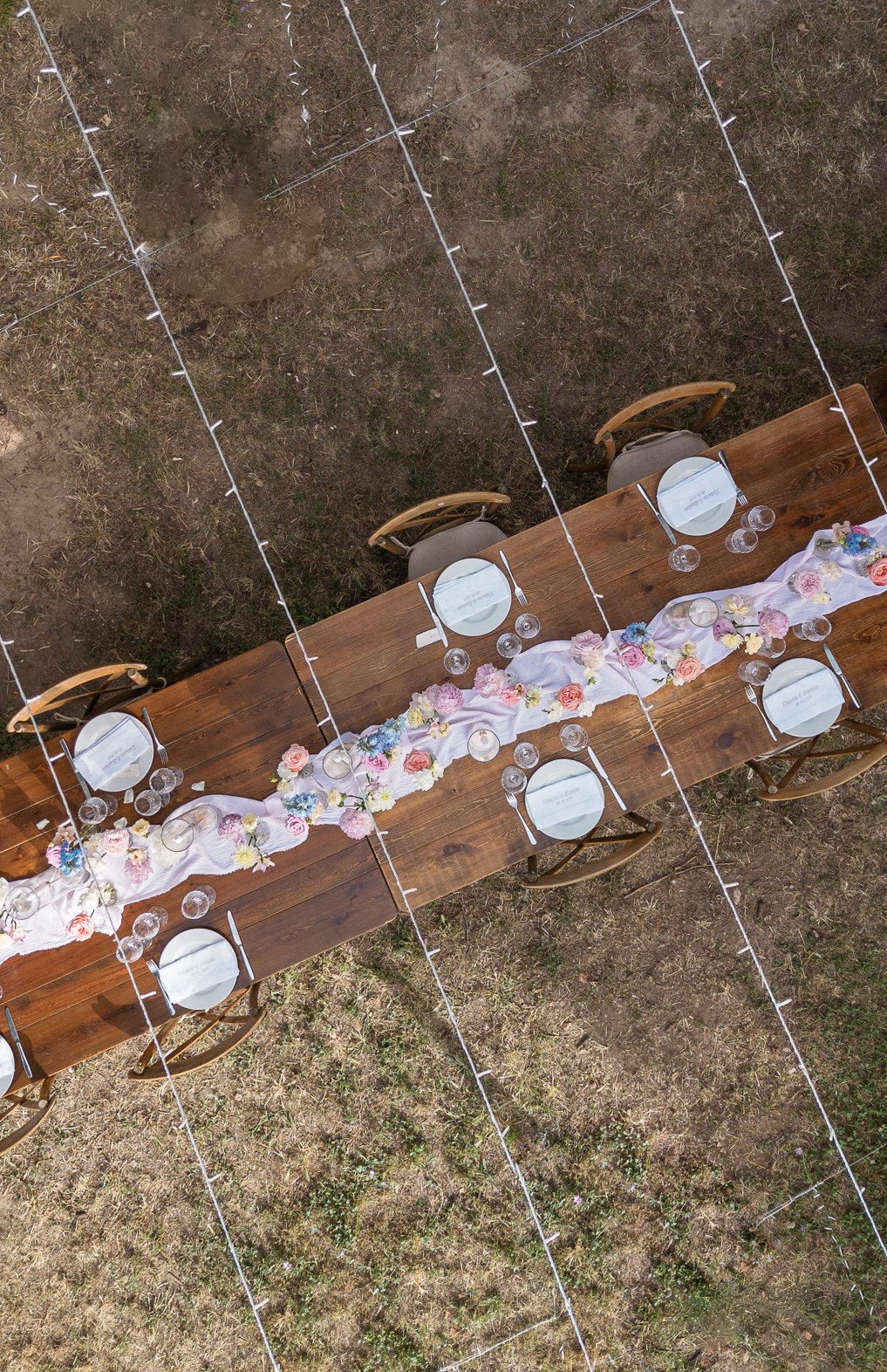 Vue aérienne d’une table en bois et de ses chaises crossback, chemin de fleurs pastel et guirlandes tendues au-dessus