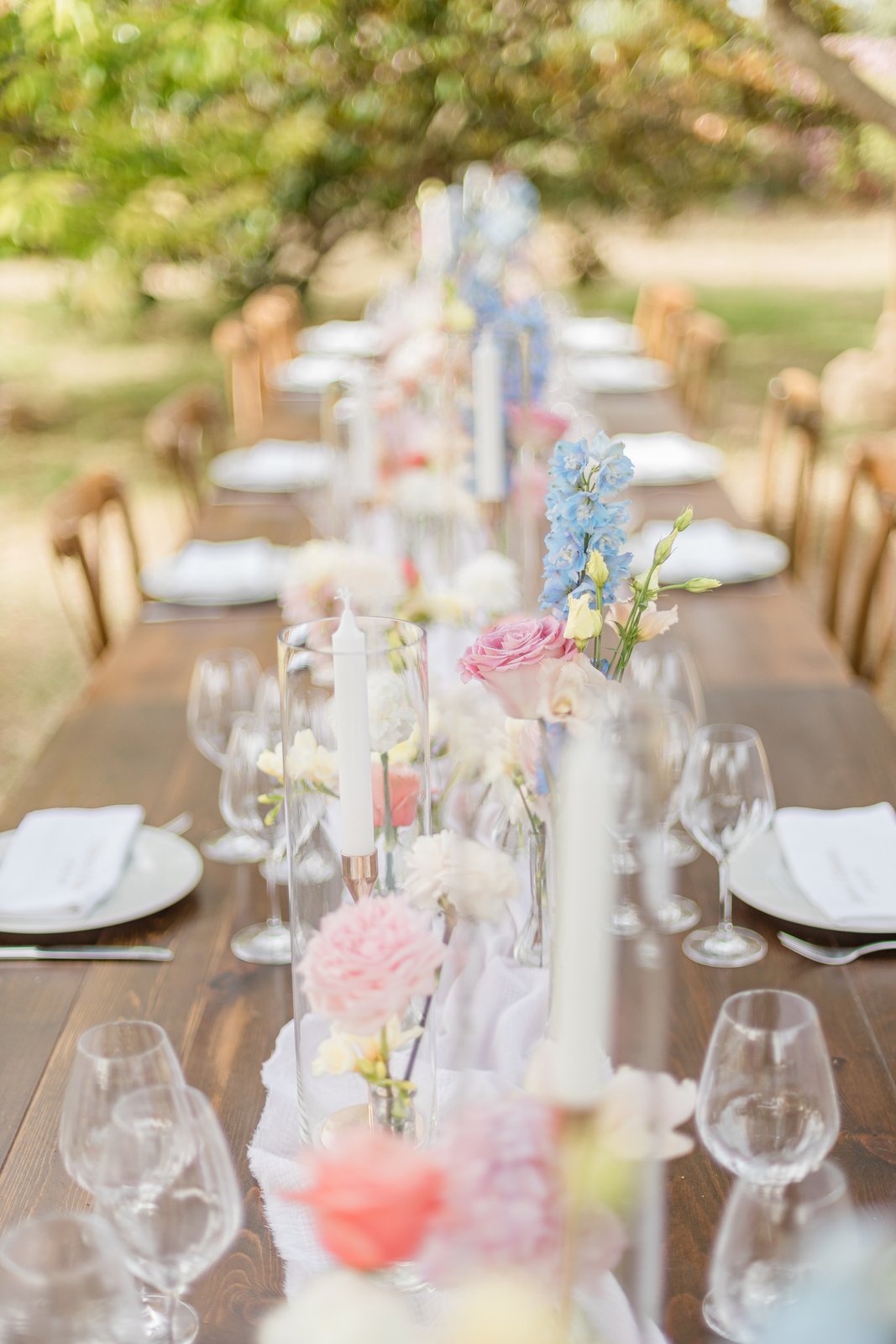 Tablée en bois dressée de jour : assiettes blanches, verres à pied, soliflores de roses et de delphiniums pastel
