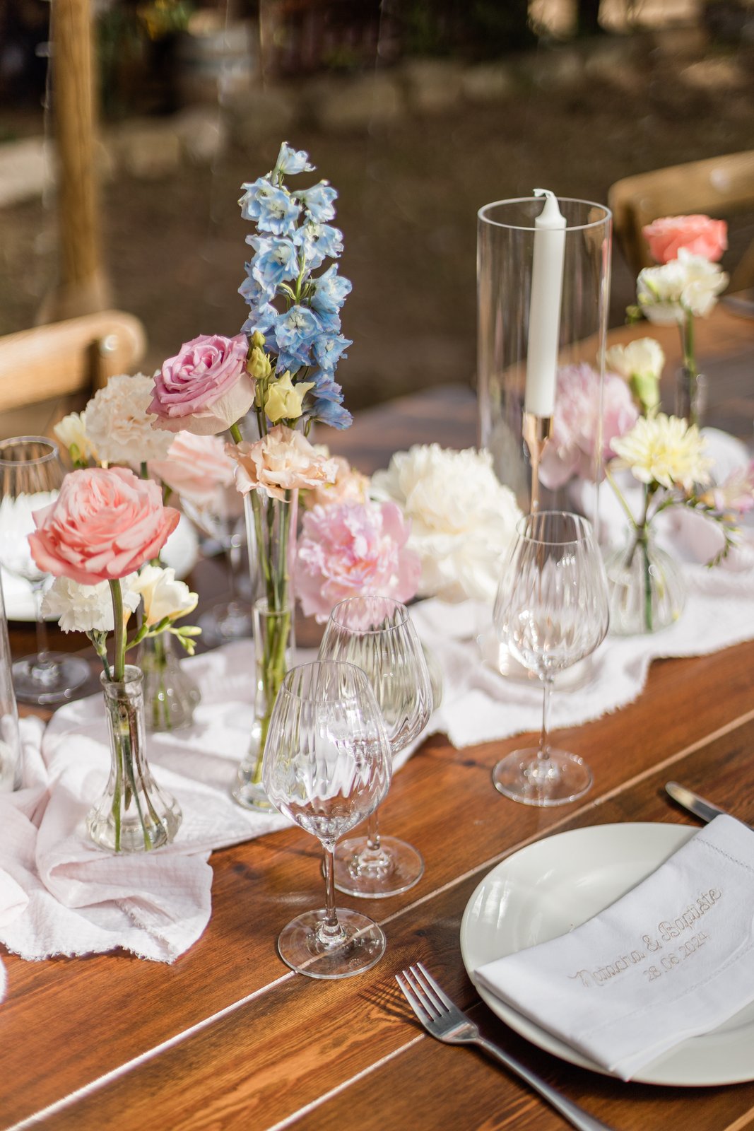 Centre de table pastel sur bois : roses, œillets et delphiniums en soliflores, bougie sous photophore, gaze blanche