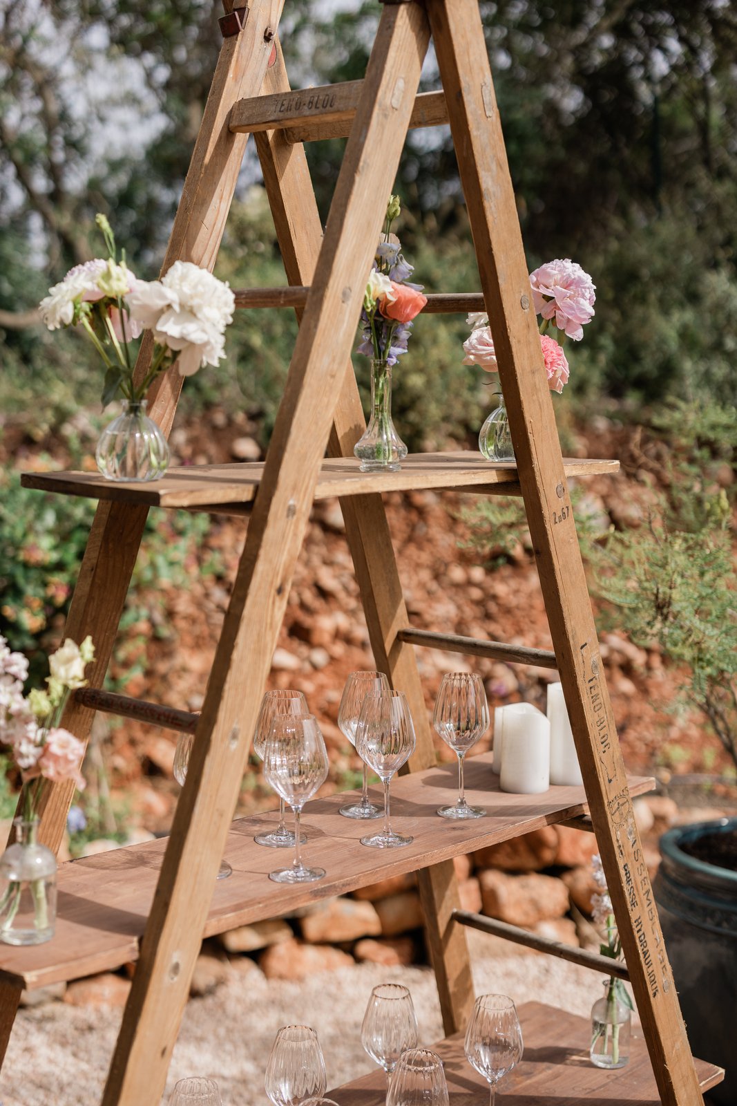 Échelle en bois à plateaux présentant verres à vin, bougies blanches et petits vases de fleurs pastel dans un jardin
