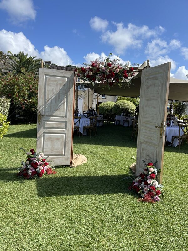 Portes de cérémonie blanches ouvertes sur la pelouse, couronnées et bordées de compositions de roses rouges et roses, tente tendue et tables en fond