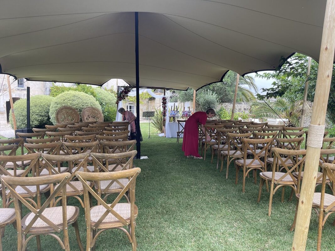Cérémonie sous une tente tendue : rangées de chaises bistrot Crossback, deux fauteuils paon en rotin et arche drapée de blanc au fond