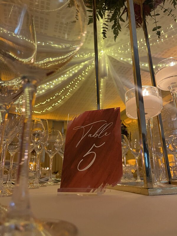 Numéro de table devant une colonne dorée et des bougies flottantes sur pied, plafond drapé de guirlandes lumineuses en reflet