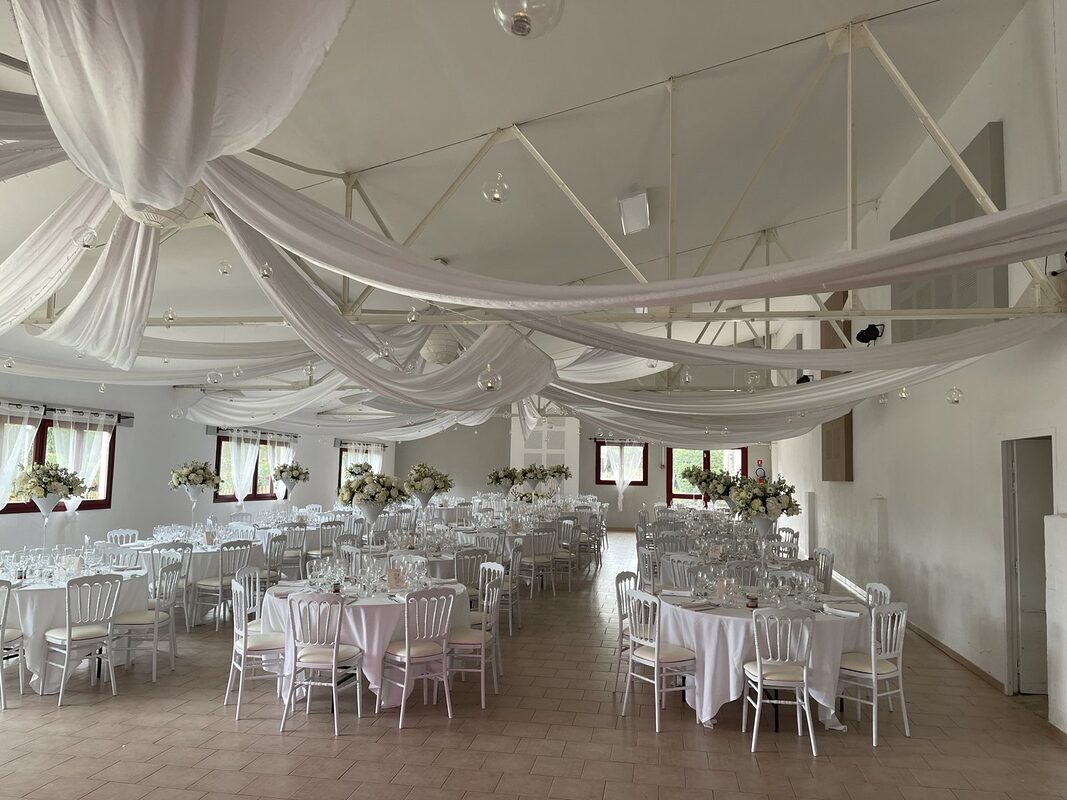 Salle de réception sous un plafond drapé de voilages blancs et de boules en verre suspendues, tables rondes nappées, chaises Napoléon III blanches et hauts vases Martini fleuris