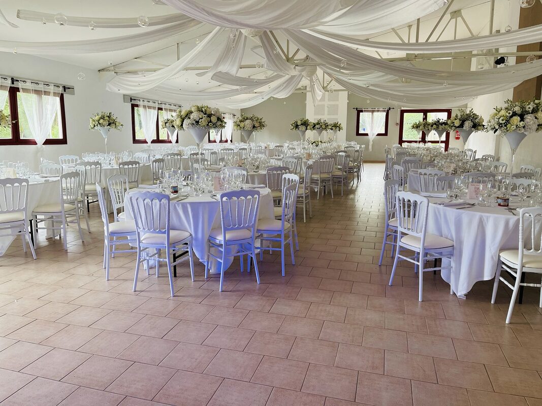 Allée centrale entre les tables rondes nappées de blanc, chaises Napoléon III blanches, centres de table fleuris sur hauts vases Martini blancs