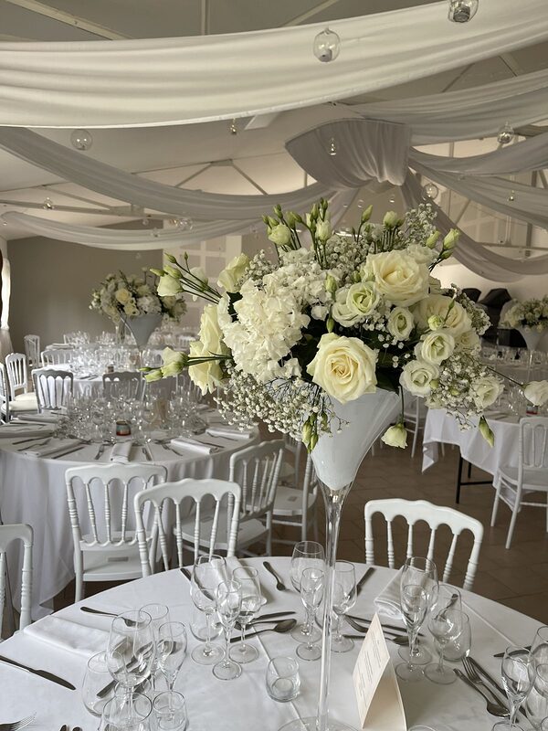 Haut vase Martini blanc garni de roses, hortensias et lisianthus blancs, au-dessus d’une table ronde dressée, voilages et boules en verre au plafond