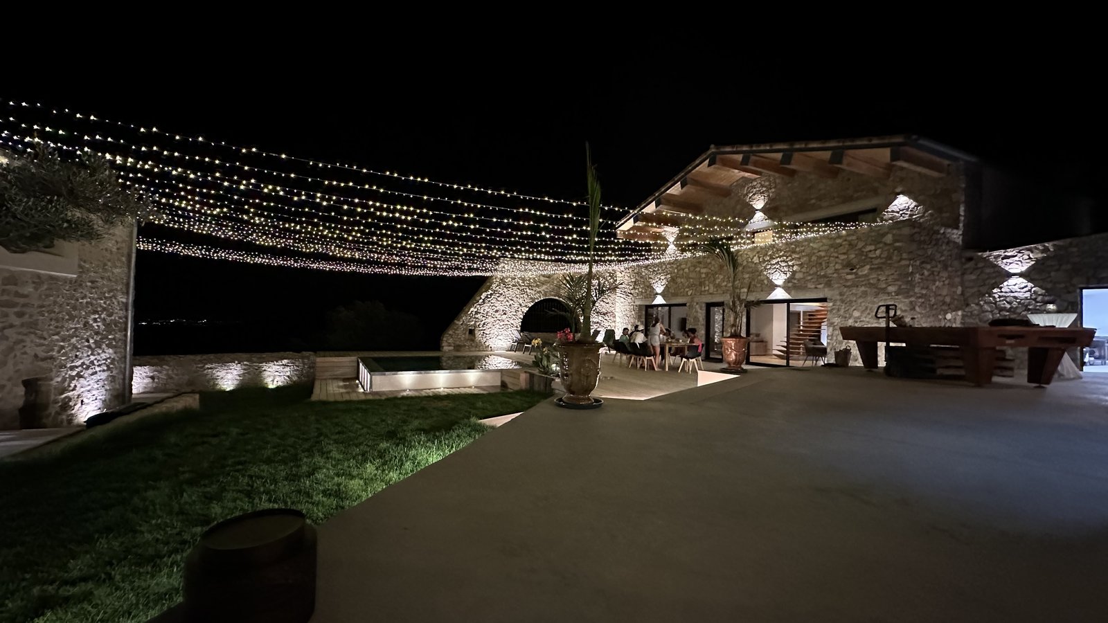 Ciel étoilé de guirlandes lumineuses tendu au-dessus de la terrasse d’un mas en pierre éclairé de nuit, pelouse et piscine