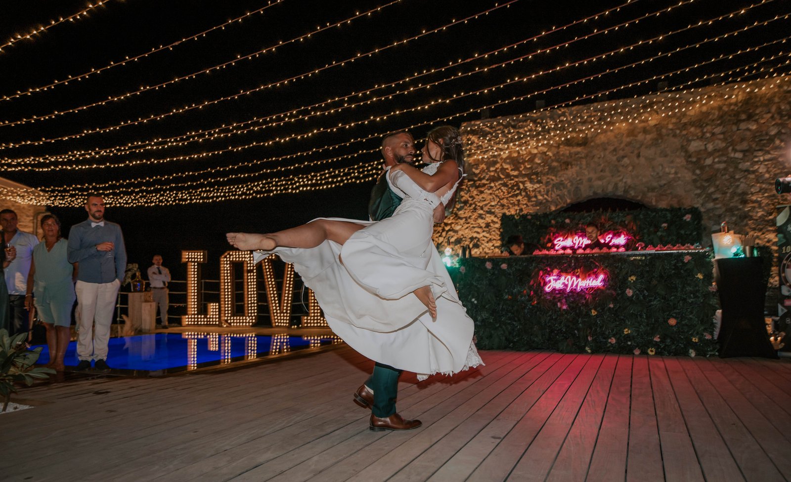 Lettres LOVE lumineuses au bord d’une piscine éclairée de bleu, sous un ciel étoilé de guirlandes, piste en bois