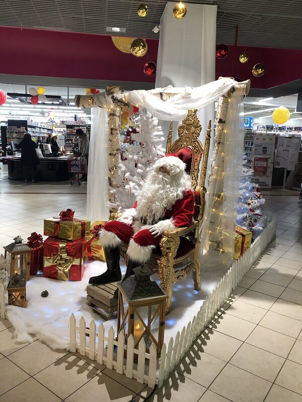 Le Père Noël sur un trône doré en velours rouge, sous une structure en troncs de bouleau drapée de voilage et de guirlandes lumineuses, lanternes et cadeaux sur la neige