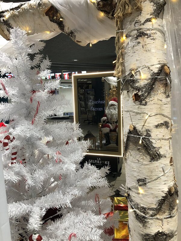 Borne photo miroir au cadre lumineux reflétant le Père Noël sur son trône, entre un sapin blanc et un tronc de bouleau