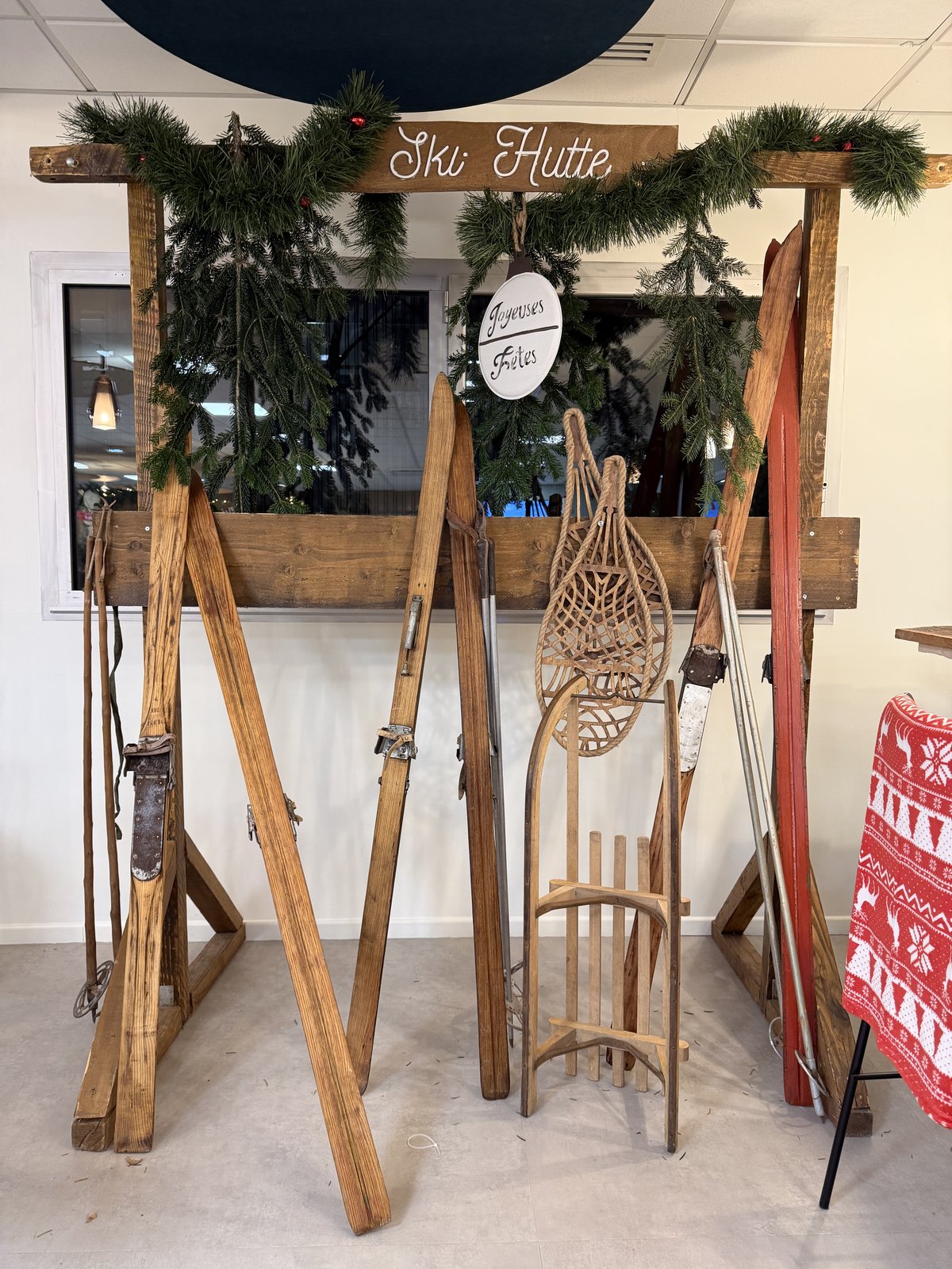 Cabane « Ski Hutte » en bois brut garnie de sapin, skis anciens, raquettes en corde et luge en bois appuyés contre la structure