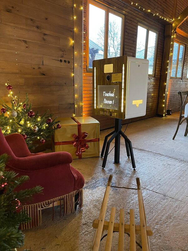 Coin photo de Noël dans une salle en bois : borne photo sur trépied, fauteuil en velours rouge à franges, cadeau doré, sapin illuminé et luge en bois