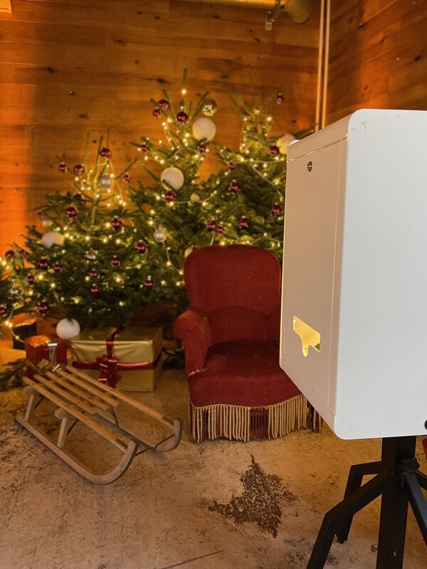 Fauteuil en velours rouge à franges et luge en bois devant les sapins illuminés, borne photo blanche au premier plan