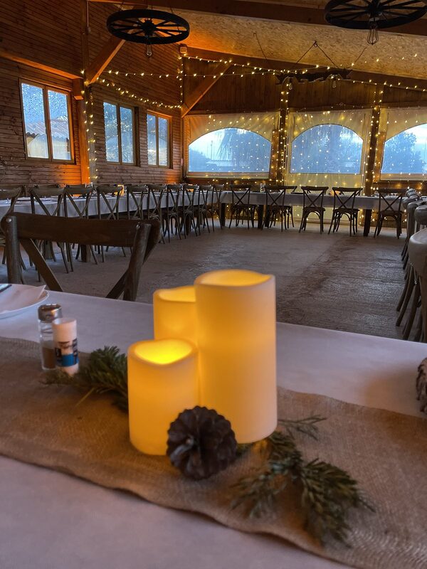 Salle du repas sous une charpente en bois : guirlandes lumineuses tendues, rideaux lumineux devant les baies, longues tables nappées de blanc et chaises en bois