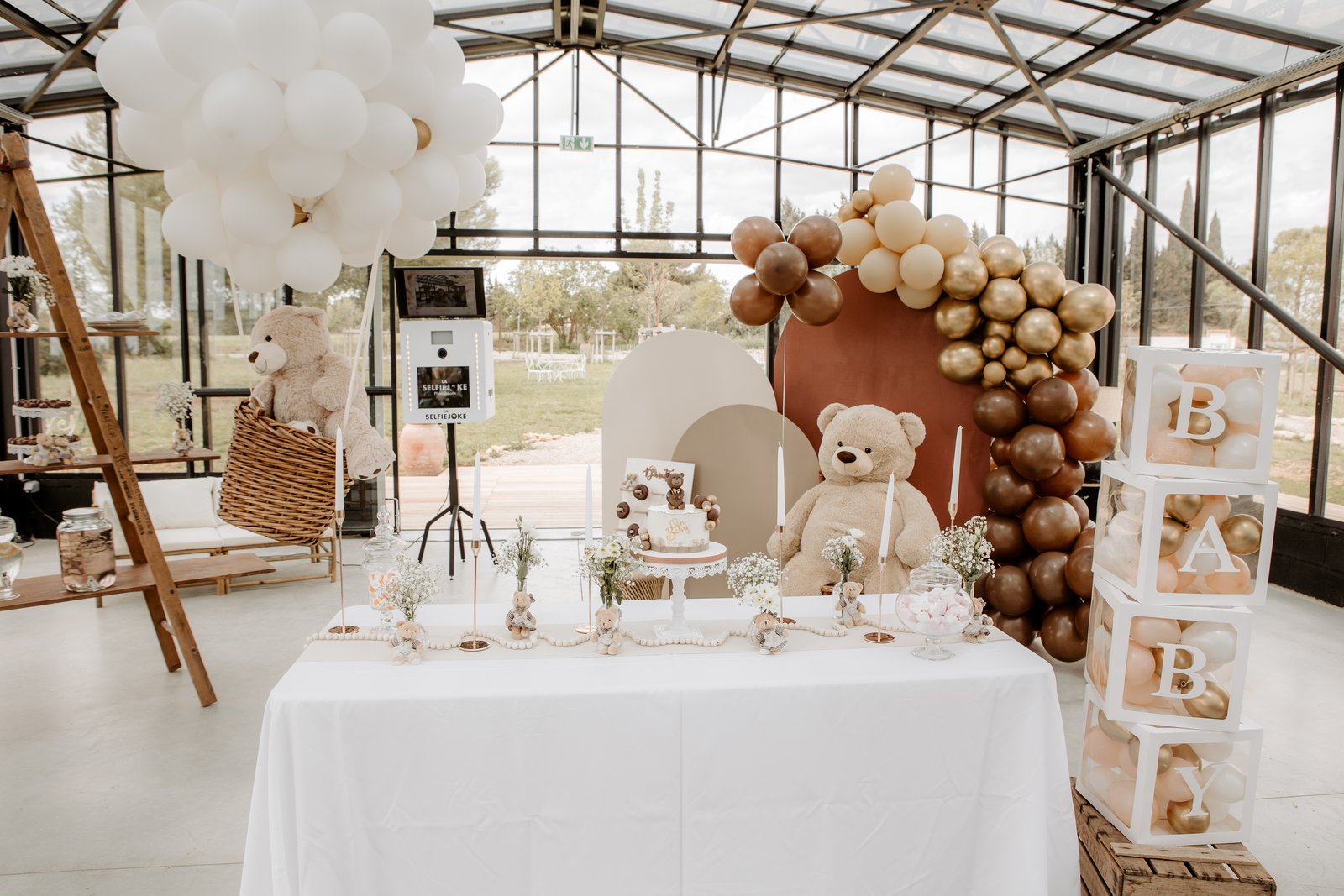 Table nappée de blanc, cubes B-A-B-Y garnis de ballons, grand nounours assis devant des arches beige et terracotta