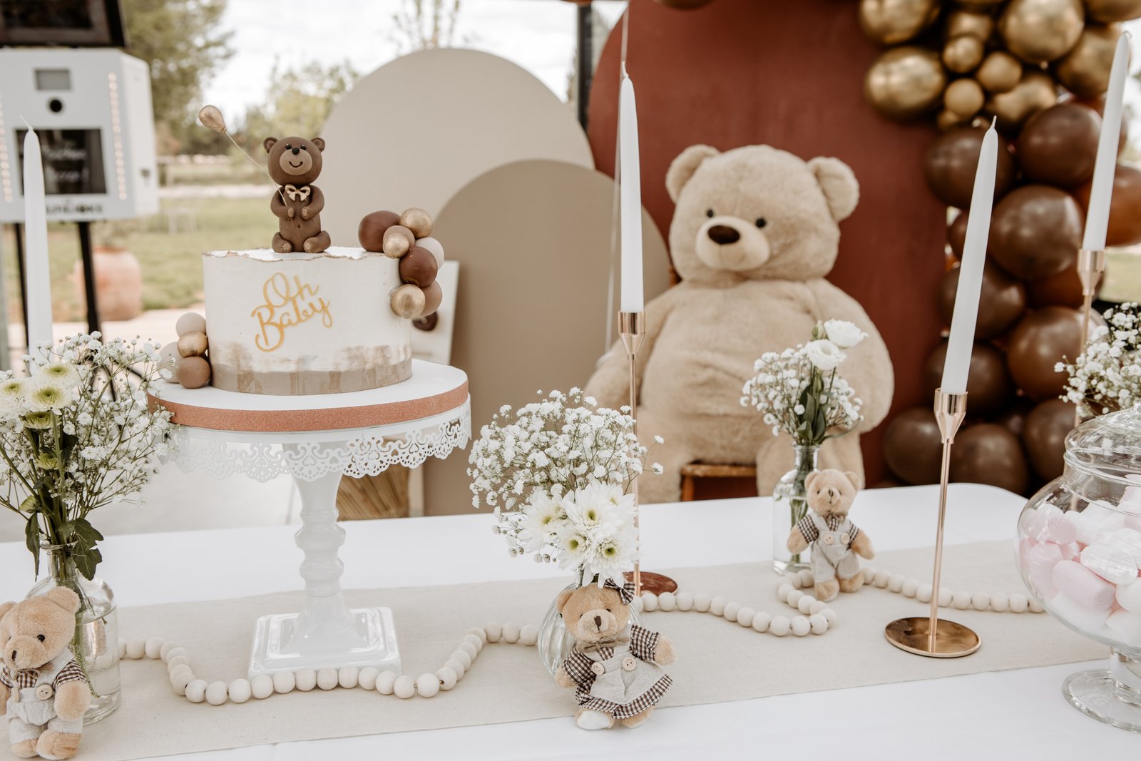 Gâteau sur support blanc ajouré, bougies blanches, gypsophile et grand nounours devant une arche de ballons chocolat