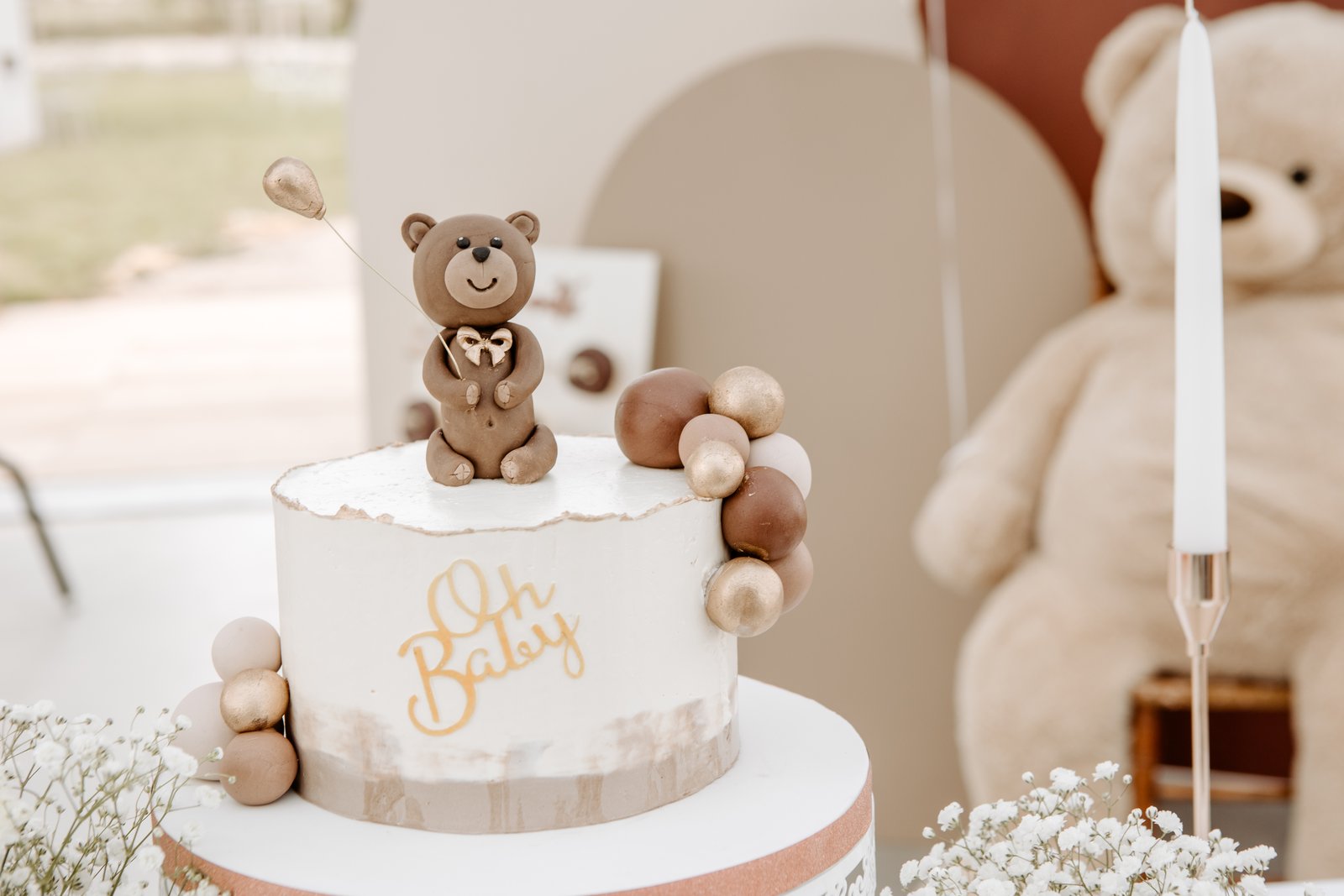 Gâteau blanc décoré de boules chocolat et dorées, grand nounours beige et bougie blanche sur bougeoir cuivré