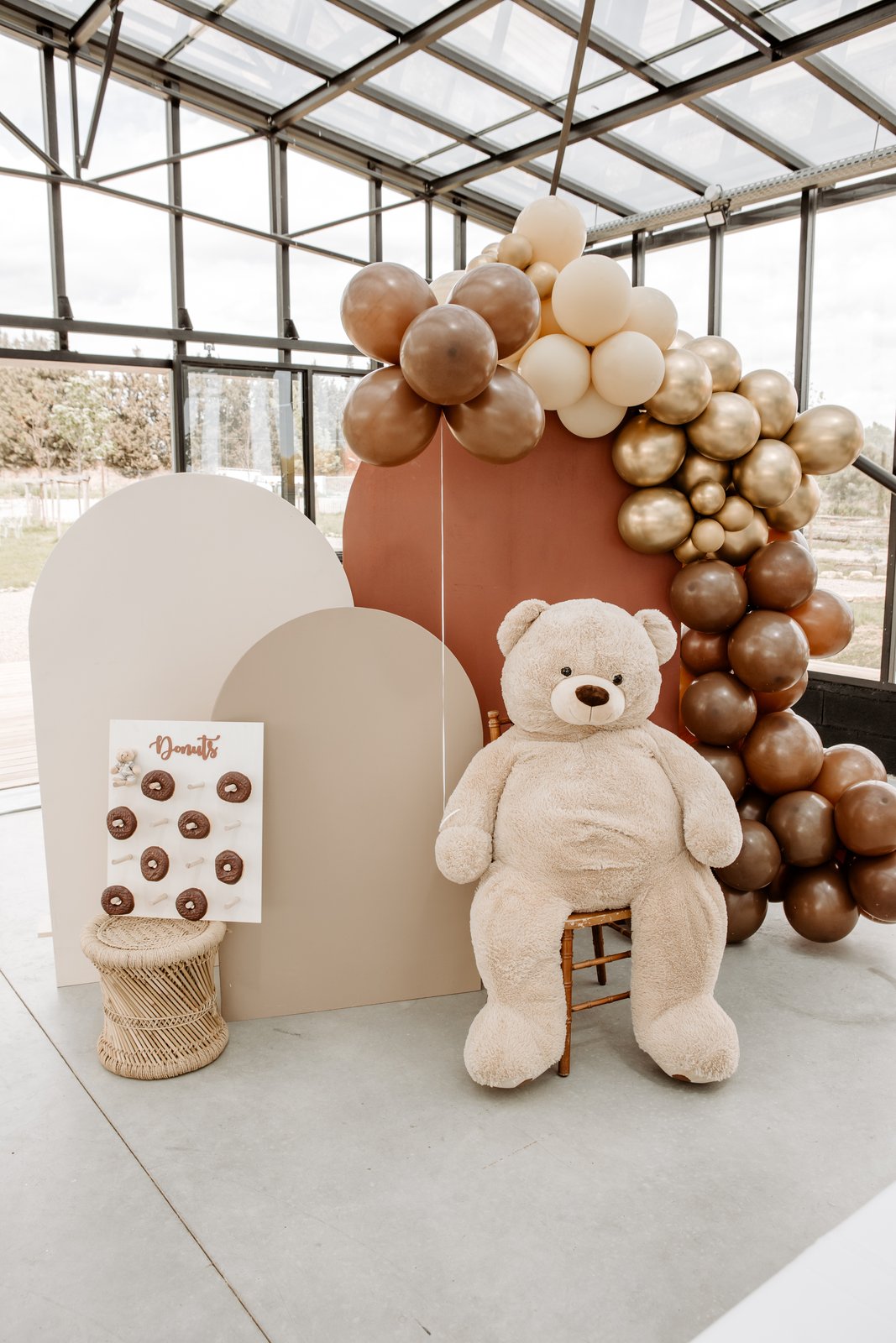 Grand nounours sur une chaise en bois, arches beige et terracotta, ballons chocolat et dorés, mur à donuts sur tabouret