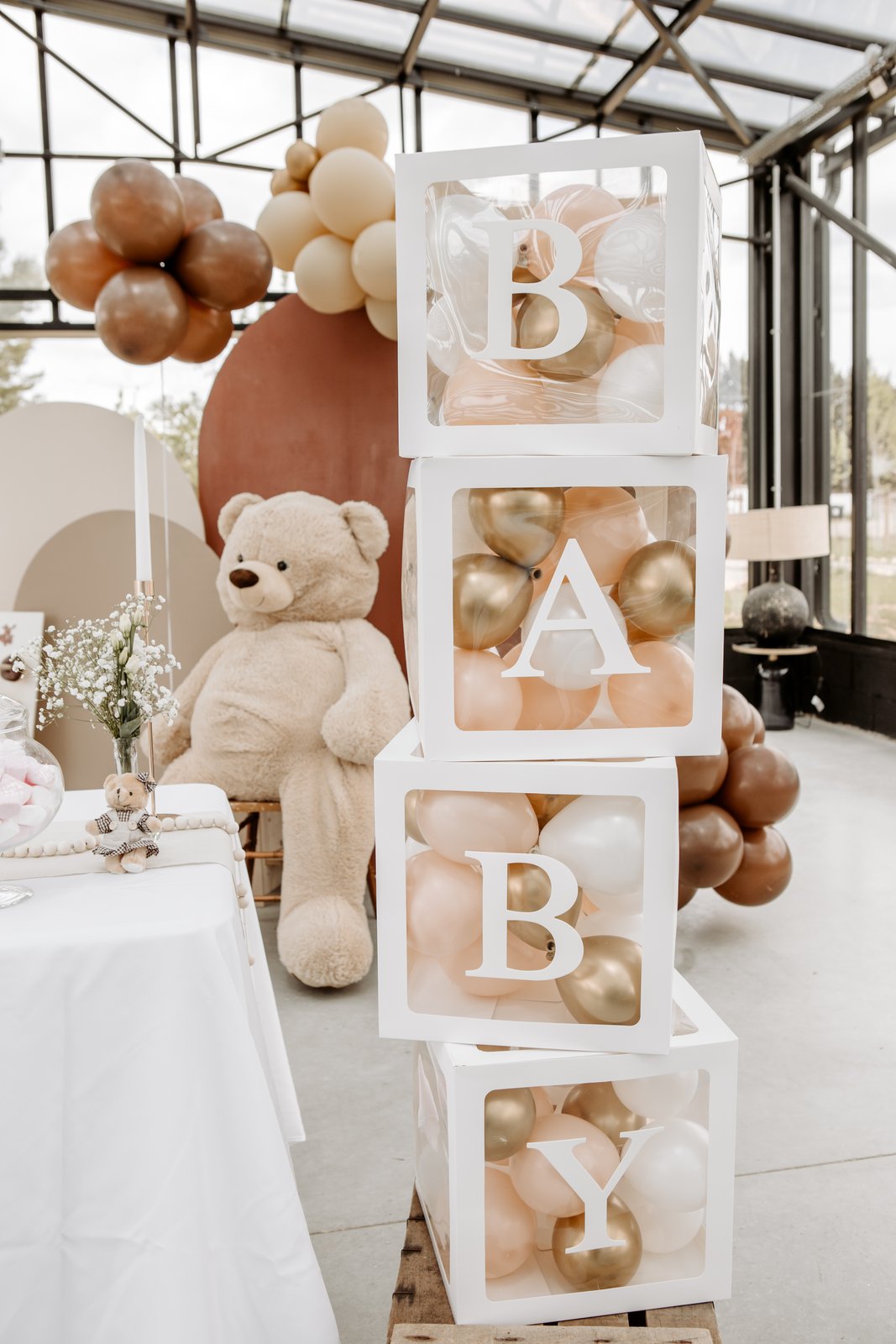 Cubes blancs B-A-B-Y garnis de ballons pêche et dorés, posés sur une caisse en bois près d’un grand nounours