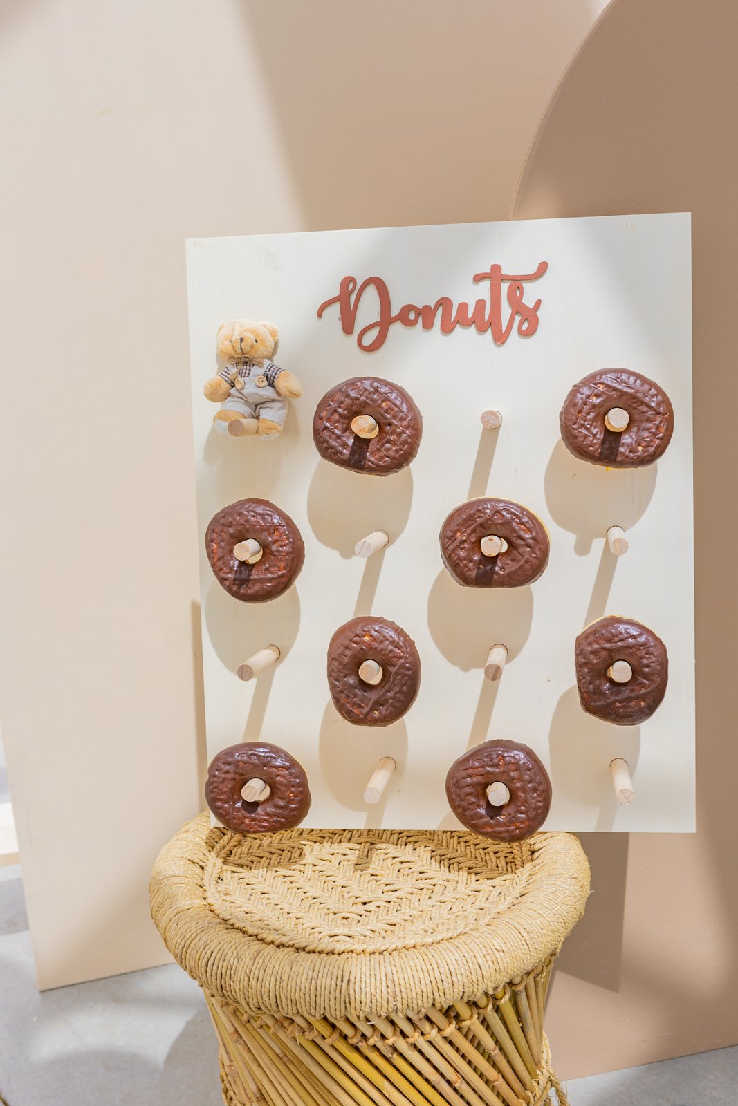 Mur à donuts blanc lettré « Donuts » et petit ourson, posé sur un tabouret en bambou et jute devant une arche beige