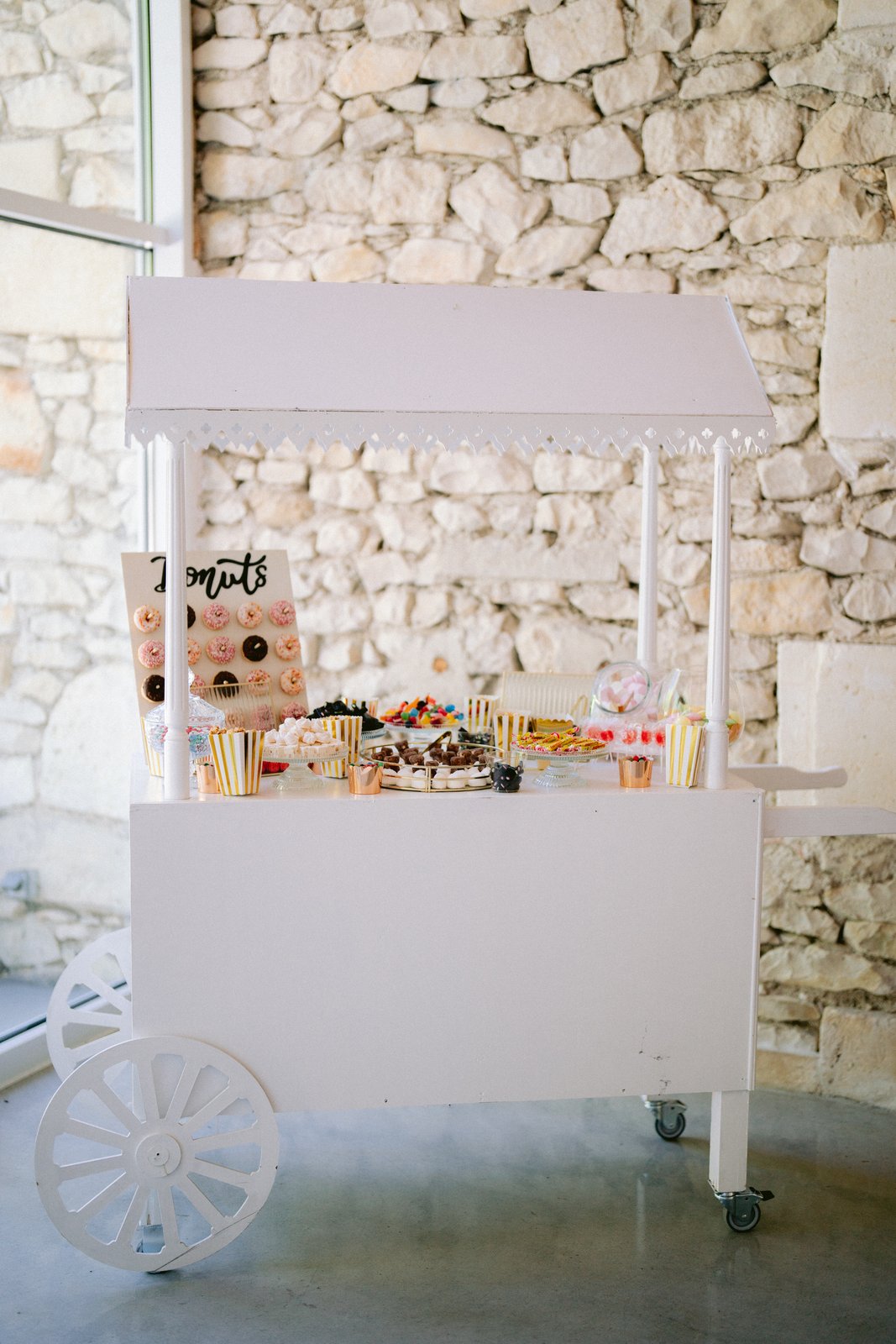 Charrette candy bar en bois blanc à roues devant un mur de pierre, garnie de bonbons, gâteaux et d’un mur à donuts