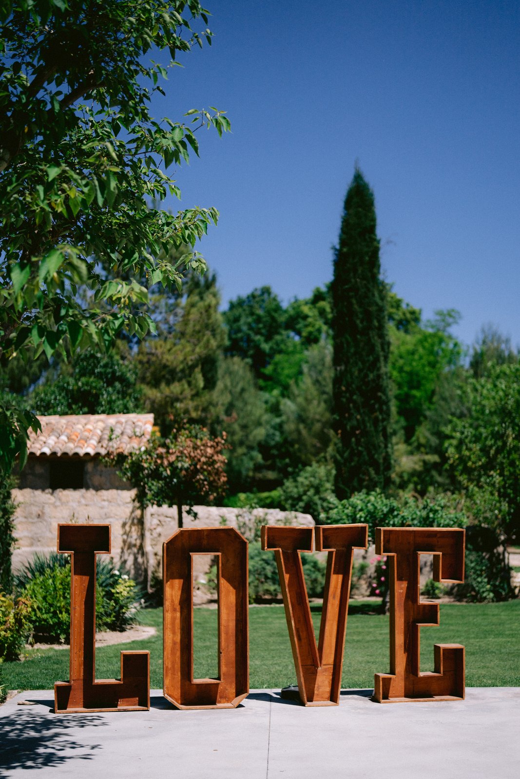 Grandes lettres LOVE en bois posées sur une terrasse, pelouse, cyprès et mas en pierre sous un ciel bleu