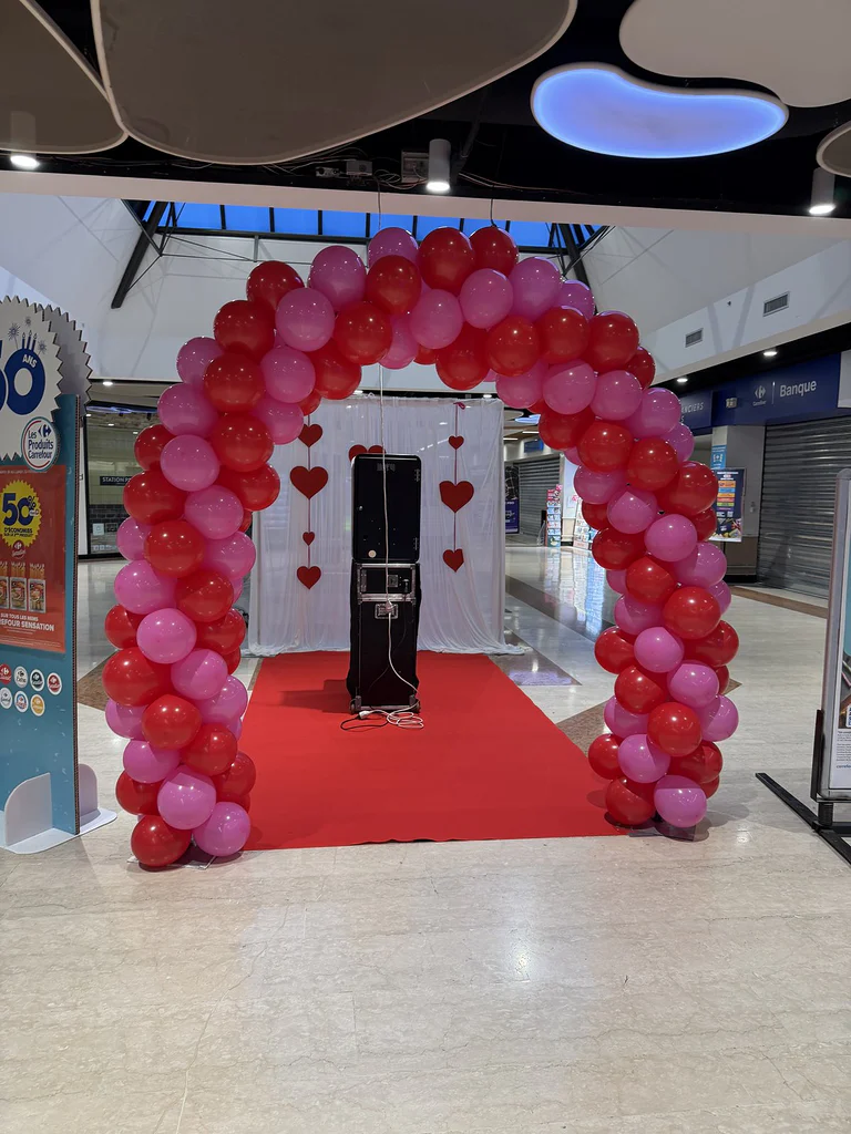 Arche de ballons rouges et roses dans une galerie marchande, tapis rouge menant à une borne photo devant un voilage à cœurs