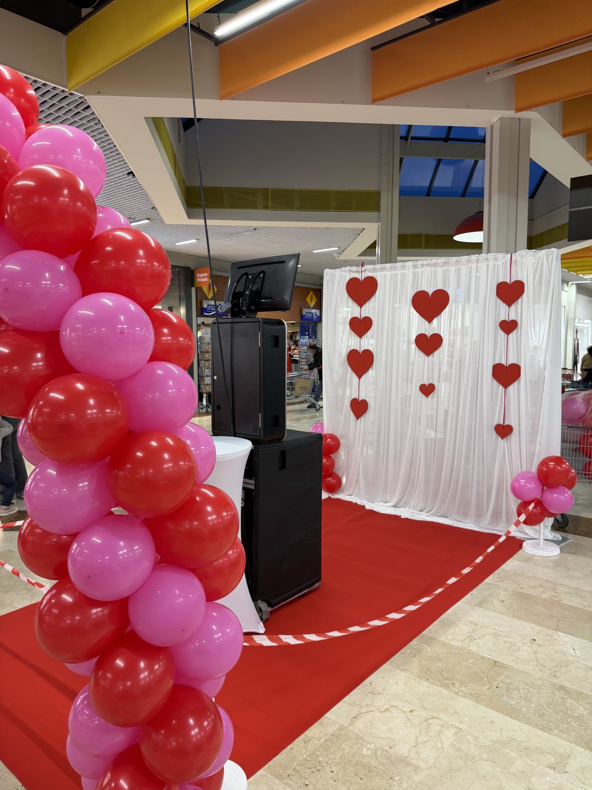 Pied d’une arche de ballons rouges et roses, borne photo noire et voilage blanc orné de cœurs rouges sur un tapis rouge