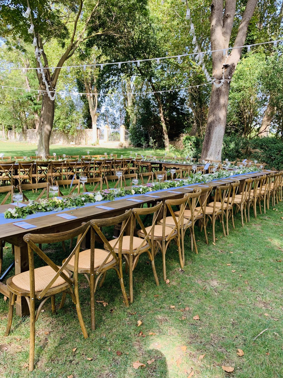 Tablée en bois avec chemin de feuillage et bougies flottantes sur pied, chaises bistrot alignées et guirlandes guinguette
