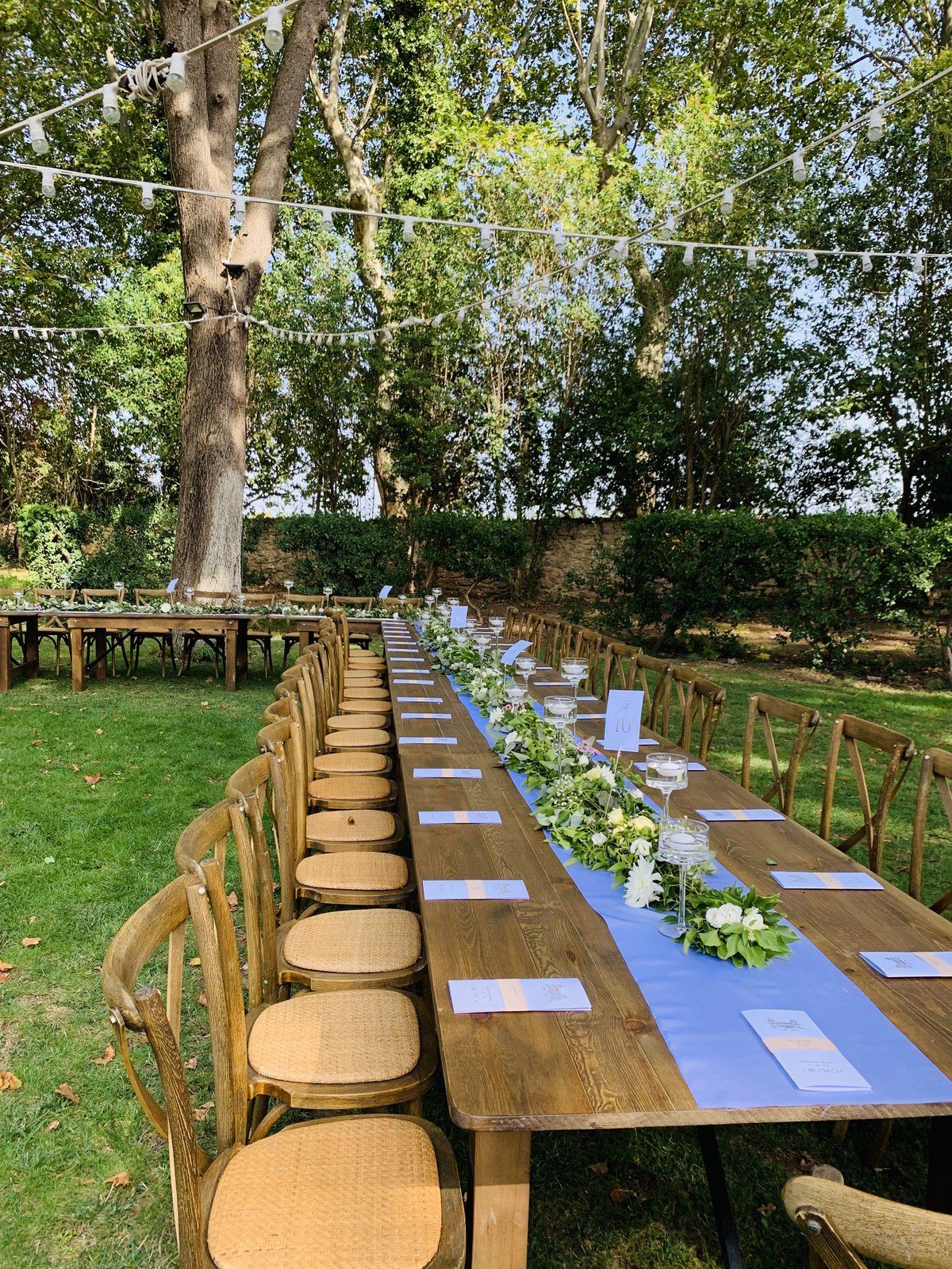 Rangée de chaises bistrot à galette effet rotin le long d’une table en bois, chemin de table bleu et guirlandes guinguette