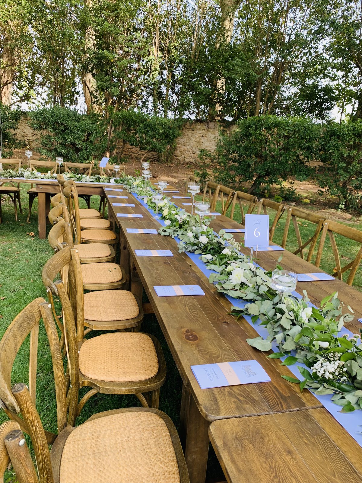 Table en bois dressée d’un chemin bleu, guirlande d’eucalyptus et de fleurs blanches, bougies flottantes sur haut pied