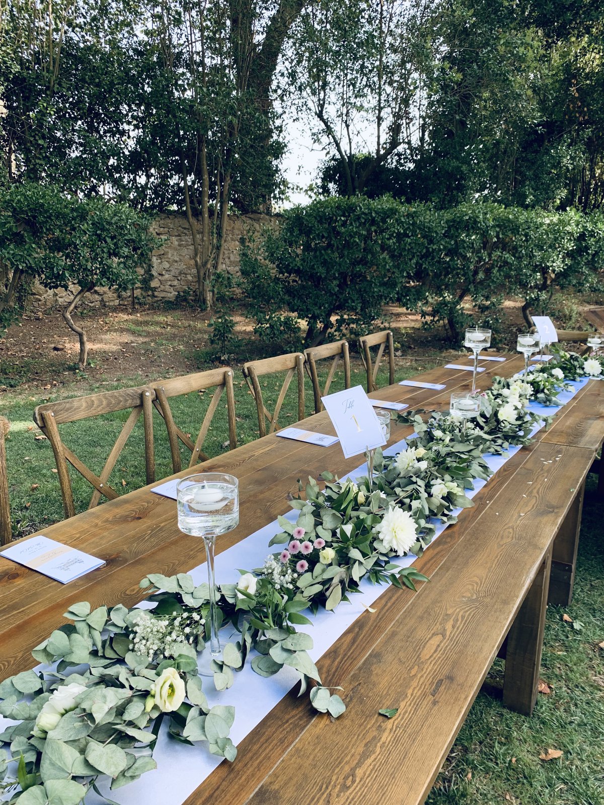 Chemin de table de feuillage et de fleurs blanches sur une table en bois, bougie flottante sur pied et chaises en bois