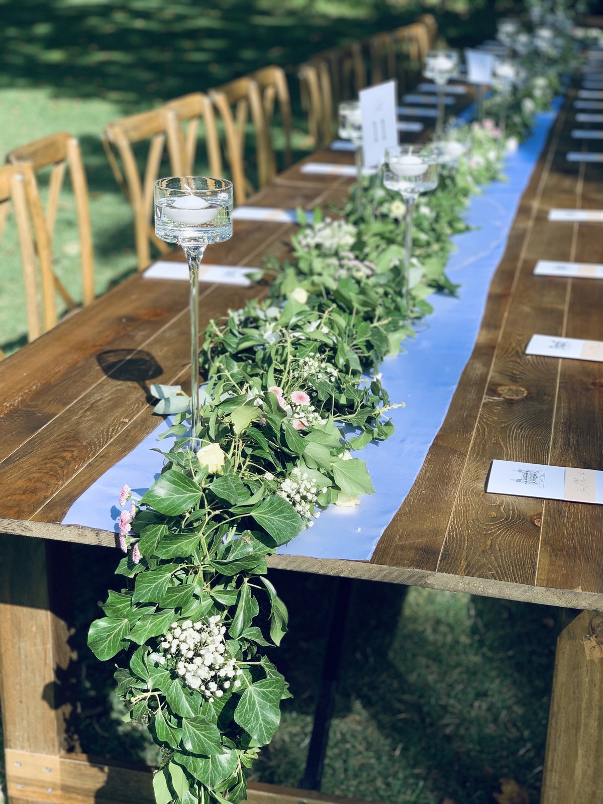 Guirlande de lierre et de gypsophile retombant d’une table en bois, chemin de table bleu et bougies flottantes sur pied