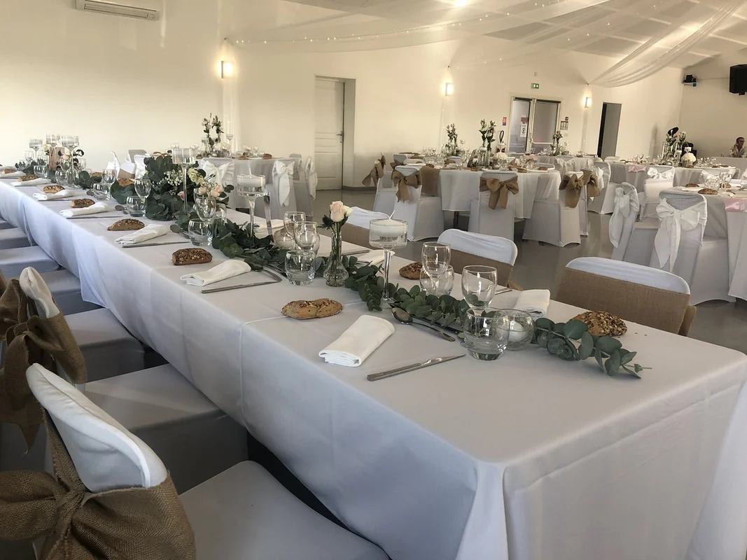 Salle de réception au plafond drapé de voile blanc et de guirlandes LED, tables rondes nappées de blanc, chaises housées aux nœuds de jute et de satin