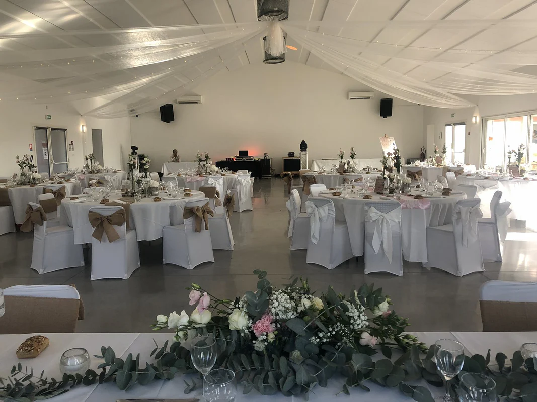 Vue de la table d’honneur sur la salle : guirlande d’eucalyptus et composition florale, tables rondes aux chaises housées, plafond drapé
