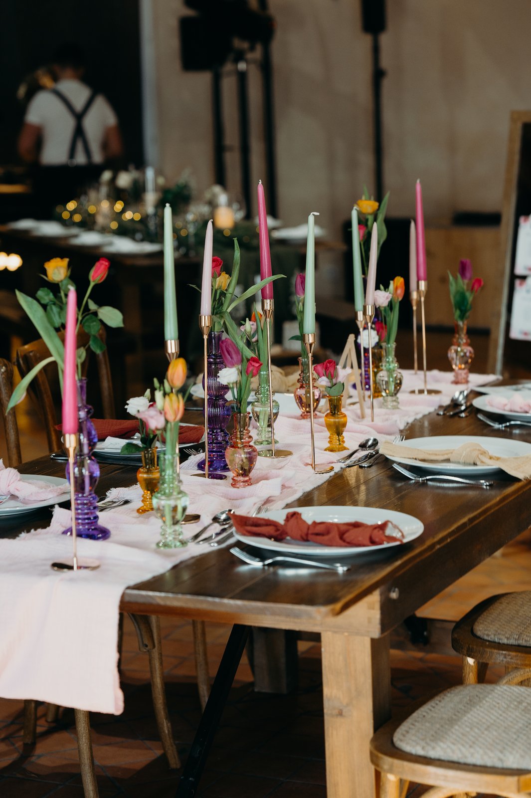 Tablée en bois dressée d’assiettes blanches, vases en verre violet, ambre et vert, bougies fines sur chandeliers dorés