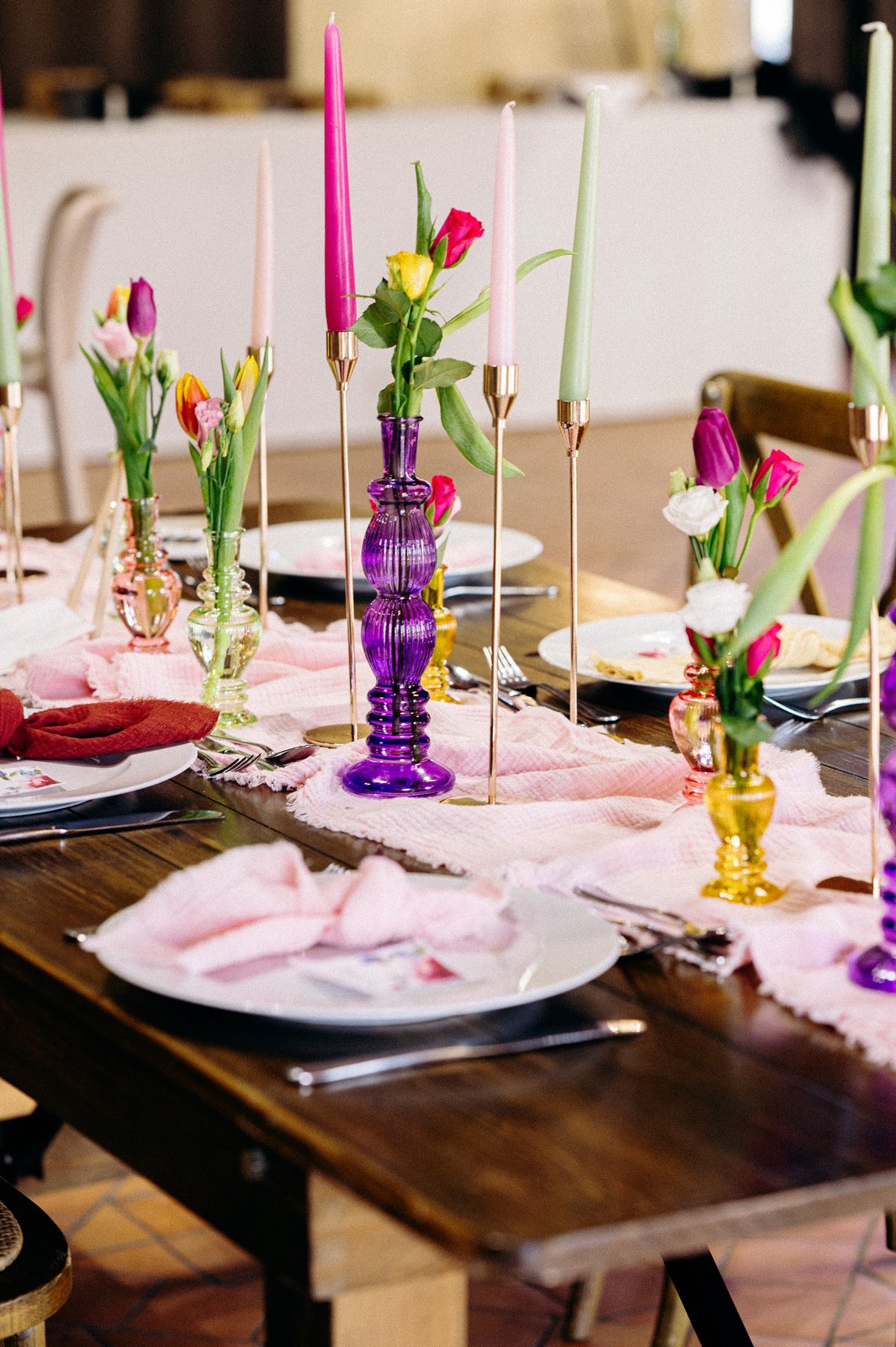 Assiettes et serviettes roses sur une table en bois, vases en verre violet et ambre garnis de tulipes, bougies roses